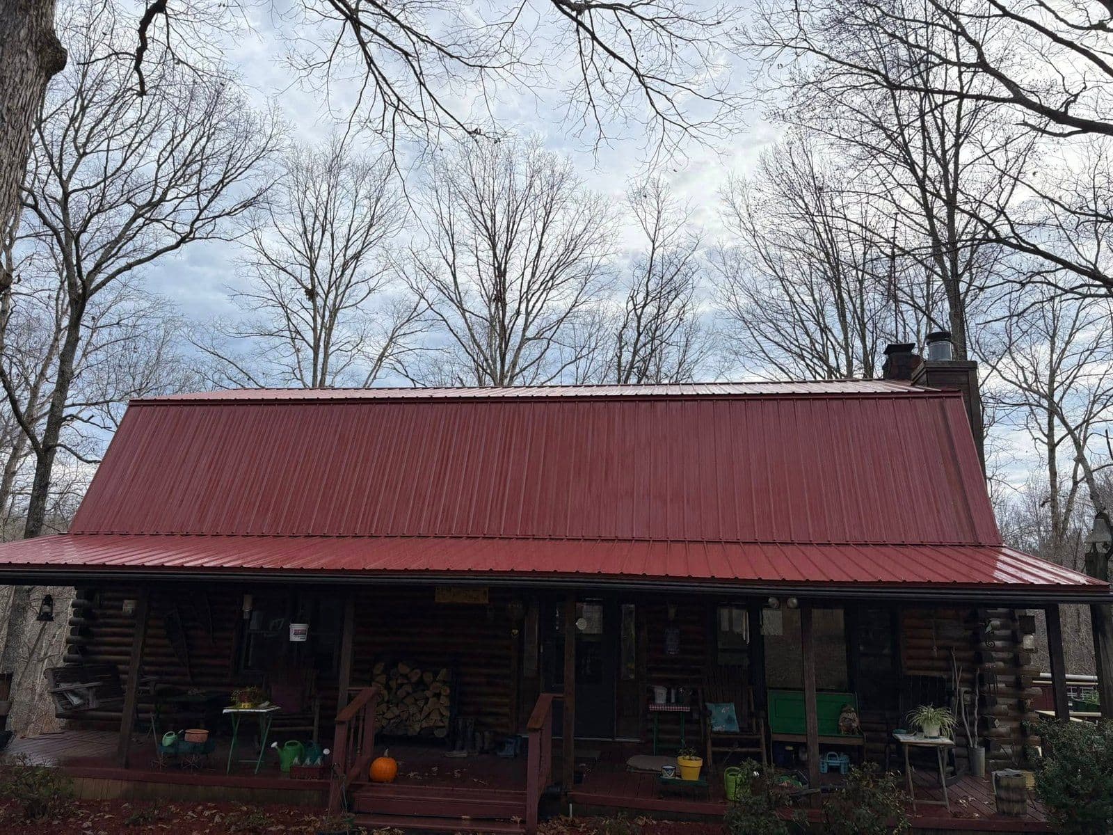 Log cabin with red metal roof surrounded by bare trees under a cloudy sky.
