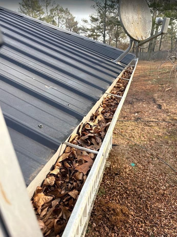 Clogged gutters filled with leaves on a metal roof, outdoor setting with trees.